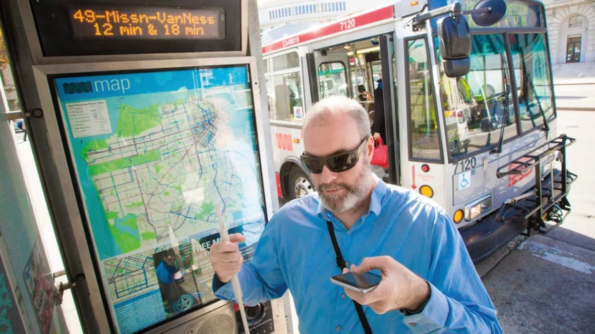 Slep moški, s sončnimi očali, belo palico v eni roki in mobilnim telefonom v drugi roki. Stoji pred veliko karto javnega potniškega prometa, v ozadju vidimo tudi avtobus.